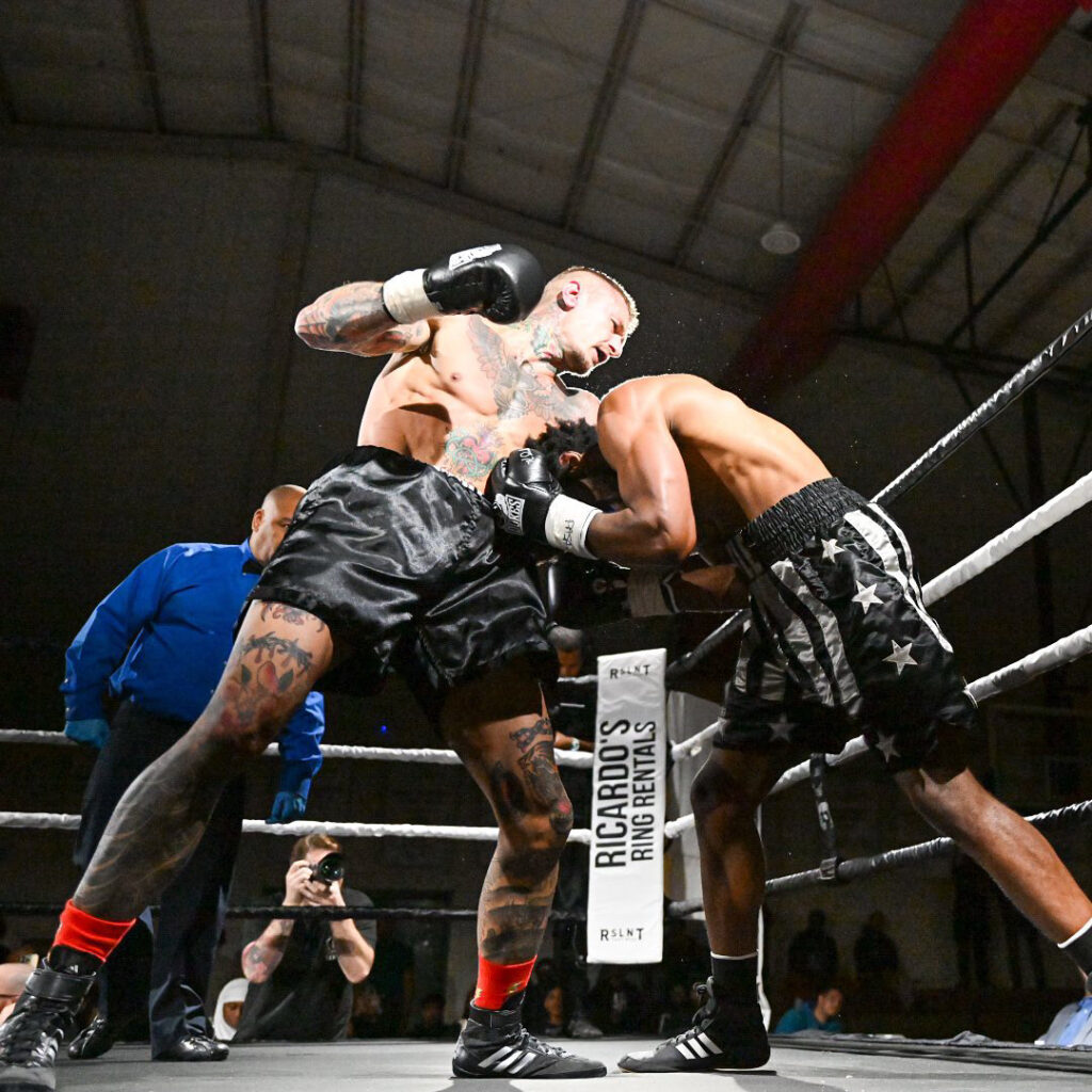 Boxers fight in the ring. Zak Kelly stands as Milton Volter leans in while placing a jab in the stomach of kelly